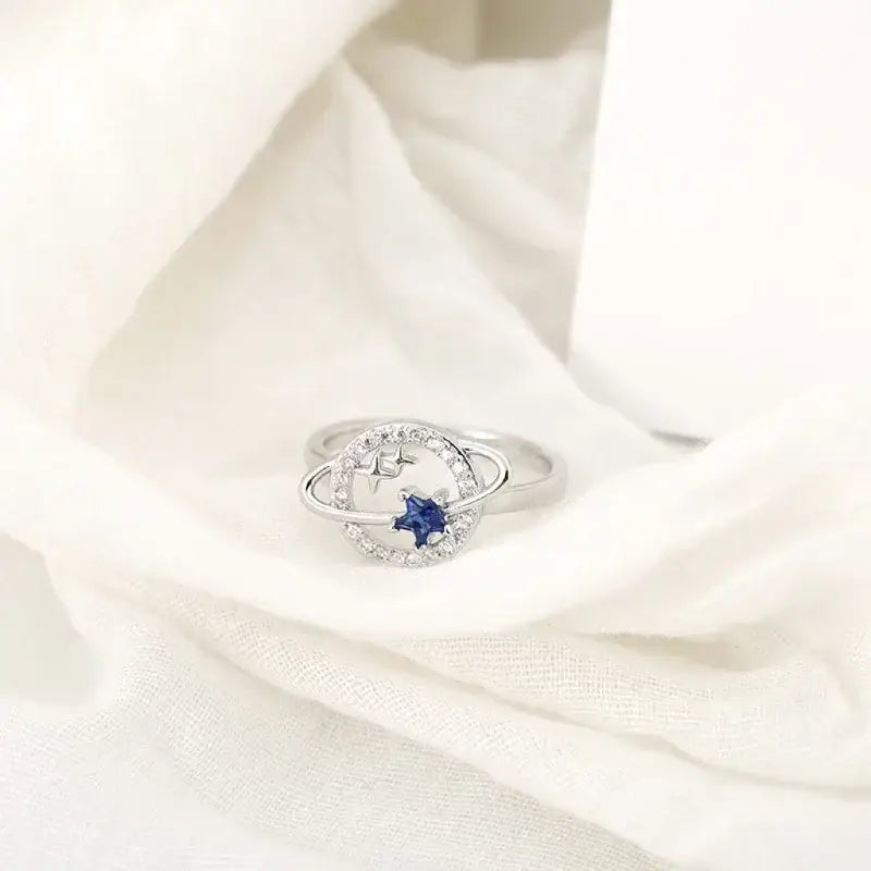 Delicado anel de prata com uma pedra preciosa azul cercada por pedras menores transparentes.