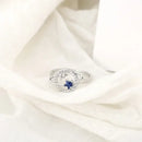 Delicado anel de prata com uma pedra preciosa azul cercada por pedras menores transparentes.