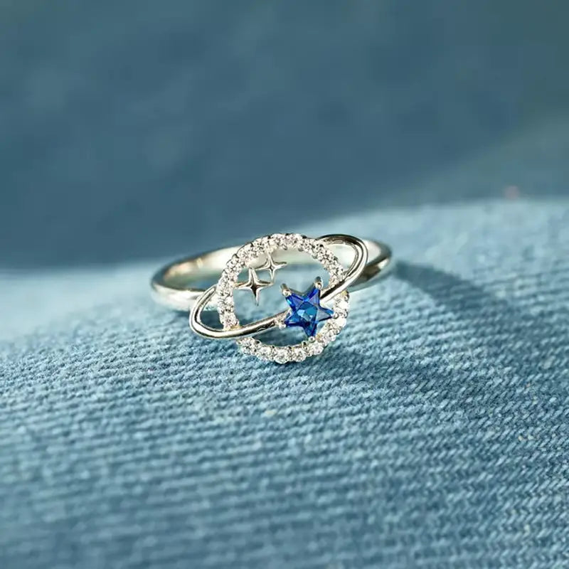 Delicado anel de prata com uma pedra preciosa azul cercada por pequenos diamantes em um design circular.